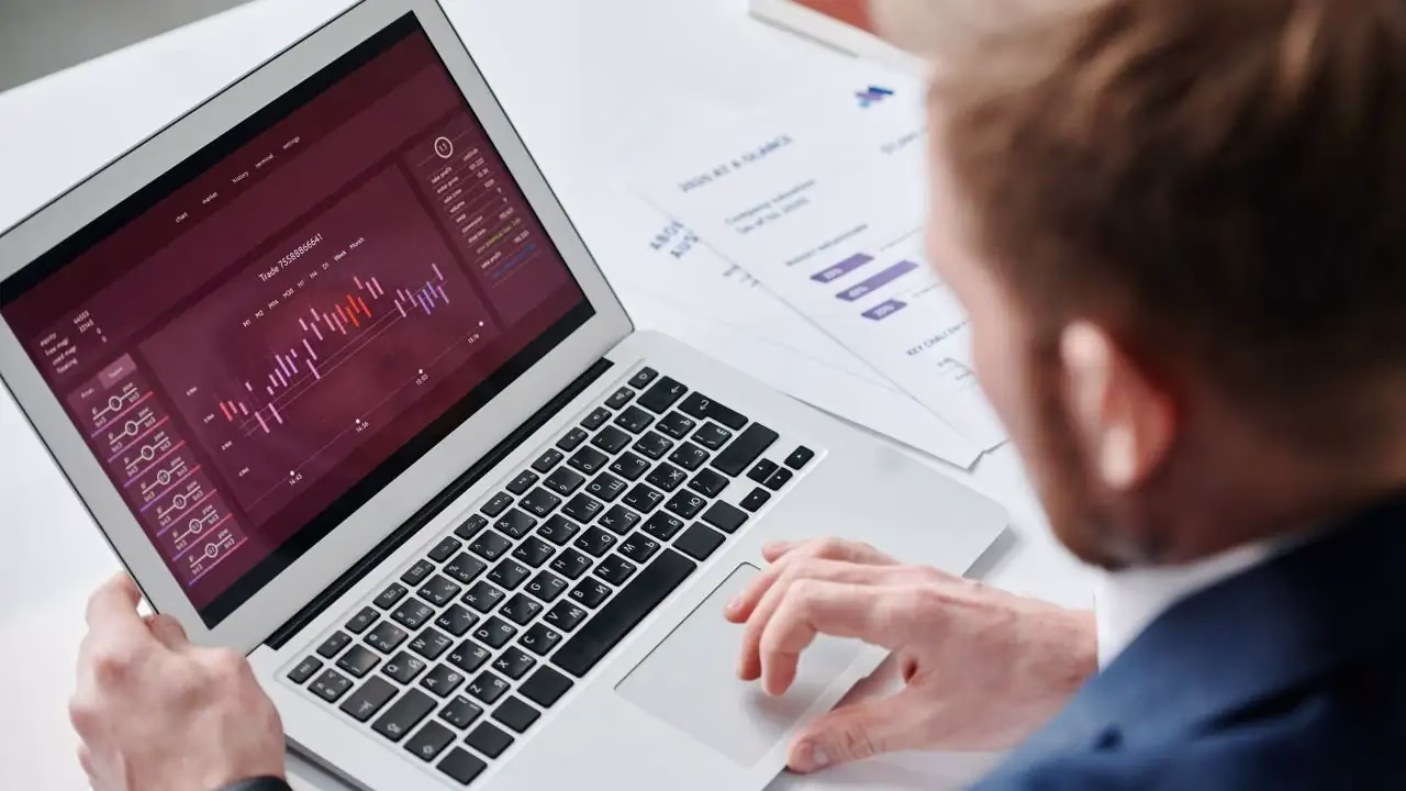 Laptop mit Diagrammen auf dem Bildschirm, daneben Unterlagen und ein Mann von hinten