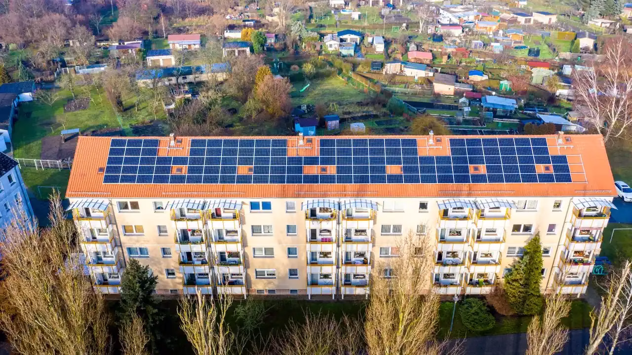 Luftaufnahme eines Mehrfamilienhauses mit Solarmodulen auf dem Dach, Kleingärten dahinter